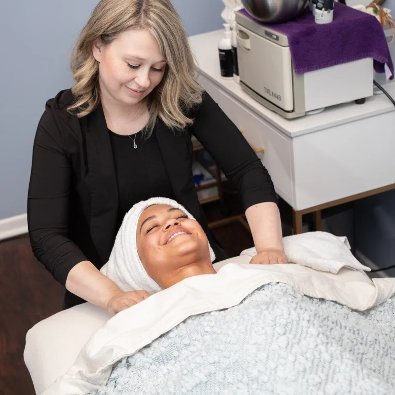 A young woman smiles while receiving a shoulder massage from a female massage therapist during a facial treatment