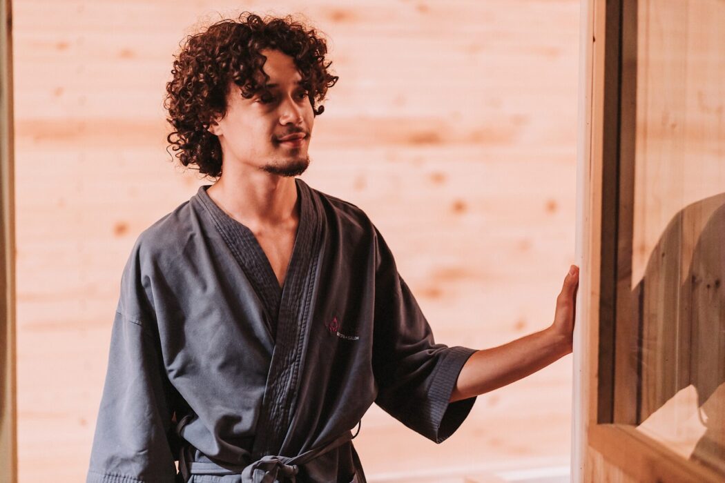 A man with curly hair stands in a sauna doorway wearing a gray bath robe